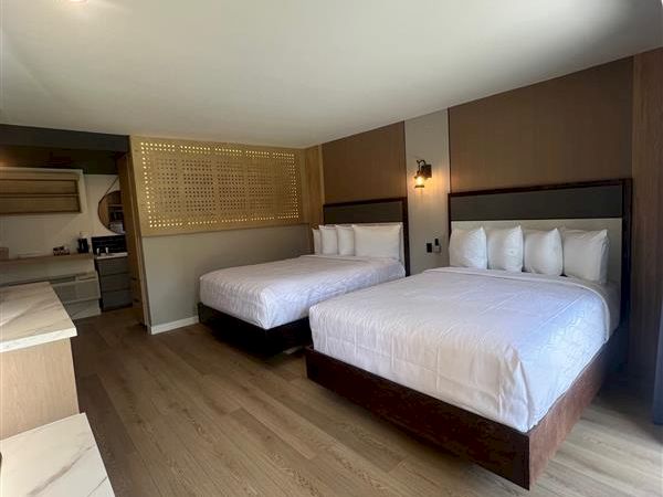 The image shows a hotel room with two double beds, wooden flooring, and a modern interior. There are lamps by the beds on the side.