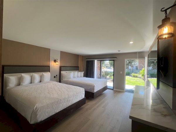 The image shows a modern hotel room with two large beds, a flat-screen TV, wooden flooring, and large windows overlooking a garden.